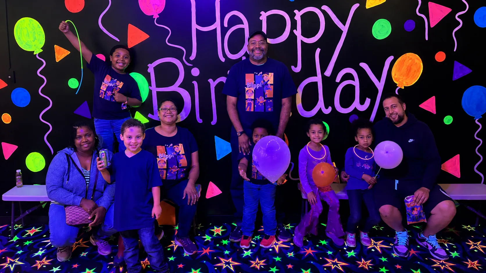 Family group posing in front of a neon Happy Birthday wall with balloons and glow decorations.