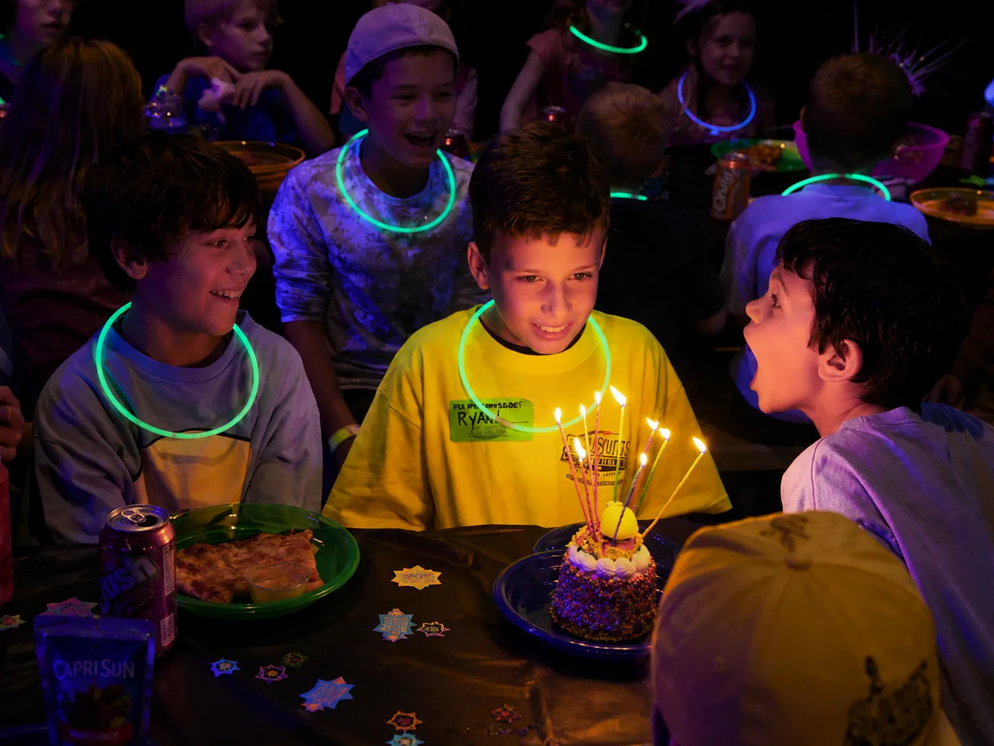 Children celebrating a birthday with cake, candles, and glow necklaces under blacklight.