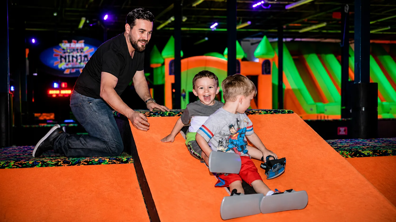 Parent helping two young children ride carpet skates down an orange slide at Fun Slides.