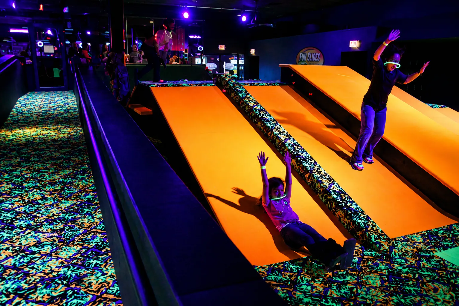 Group of kids cheering while sliding down an orange carpet slide under blacklight.