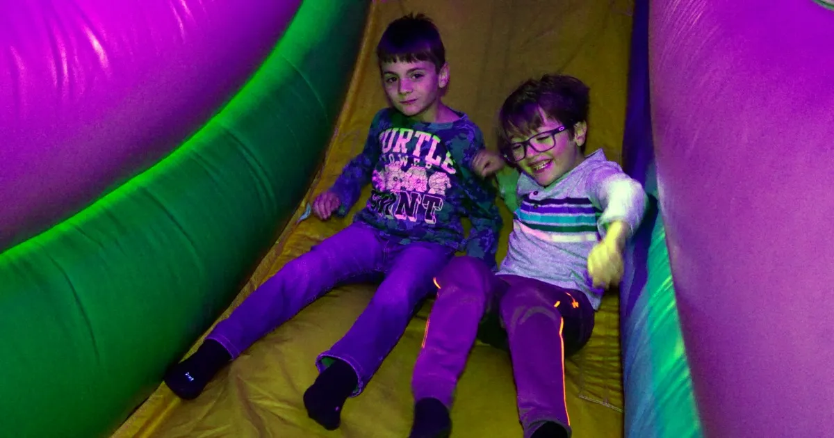 Younger kids on a bounce house while older kids skate carpeted ramps under blacklights
