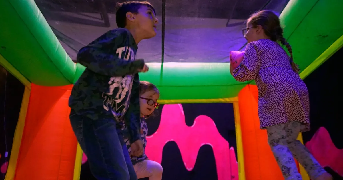 Kids of different ages skating, bouncing, and playing together inside a blacklight carpet skatepark in Pittsburgh